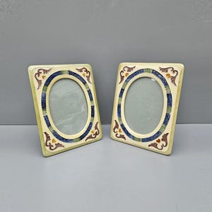 May include: Two oval picture frames with a cream background and blue, green, and orange accents. The frames are made of ceramic and have a rustic, vintage look.