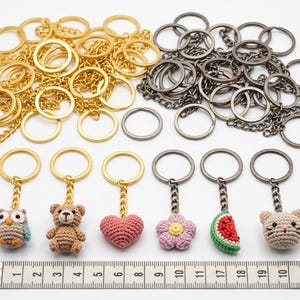May include: A collection of gold and gunmetal keychains with metal rings and chains. Six finished keychains feature crocheted charms: an owl, a teddy bear, a pink heart, a flower, a watermelon slice, and a cat head, aligned with a 12-inch ruler.