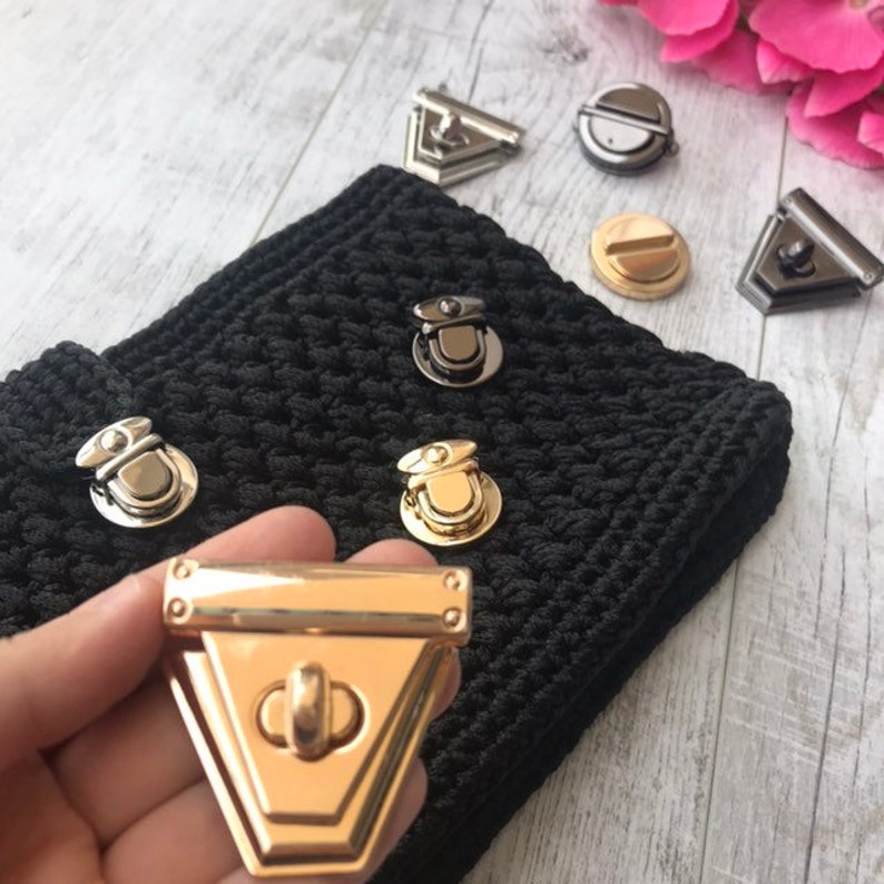 May include: Various metal purse clasps and closures in gold and silver hues, arranged on black crocheted fabric. The clasps feature diverse shapes, such as triangular and circular forms, ideal for crafting bags and accessories.