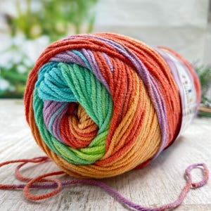 May include: A round ball of variegated yarn featuring a vibrant gradient of colors including orange, red, yellow, green, teal, and light purple. The yarn is wound in a circular pattern with a paper label attached to the side and loose strands.