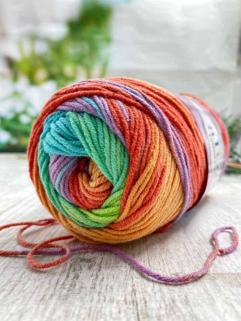 May include: A round ball of variegated yarn featuring a vibrant gradient of colors including orange, red, yellow, green, teal, and light purple. The yarn is wound in a circular pattern with a paper label attached to the side and loose strands.