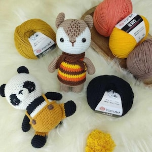 Puede incluir: Juguetes de ganchillo hechos a mano, incluyendo un panda y una criatura parecida a un ciervo, junto con ovillos de lana en varios colores. Los colores de la lana incluyen amarillo, naranja, marr&oacute;n y negro. Los juguetes est&aacute;n sobre una superficie blanca y esponjosa.