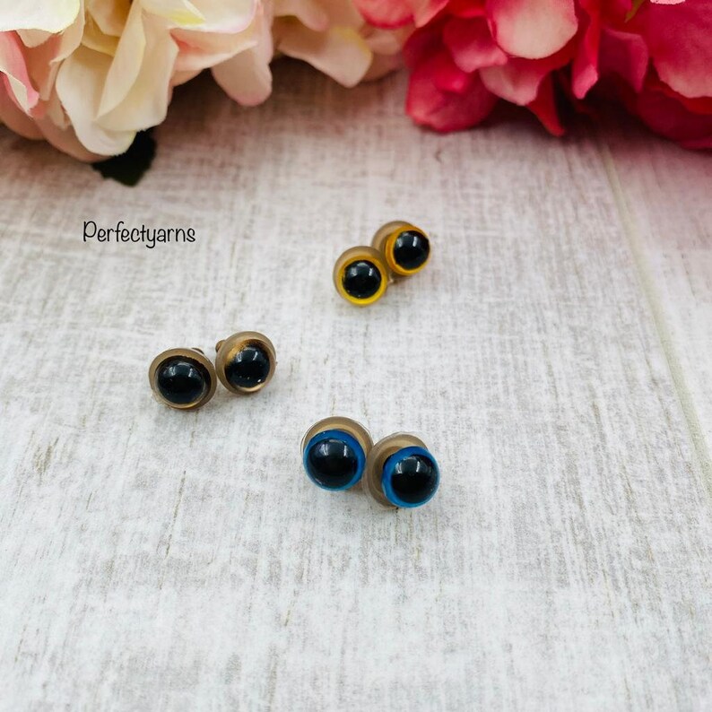 May include: Three pairs of round safety eyes for crafts, each featuring a black pupil and a colored iris. One pair has yellow irises, one pair has brown irises, and one pair has blue irises. The text Perfectyarns is visible on the left side.