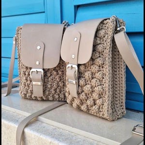 May include: Two beige crochet crossbody bags with leather flaps and silver buckles. The bags have a textured, bobble-like pattern and adjustable straps. The background is a blue shutter.