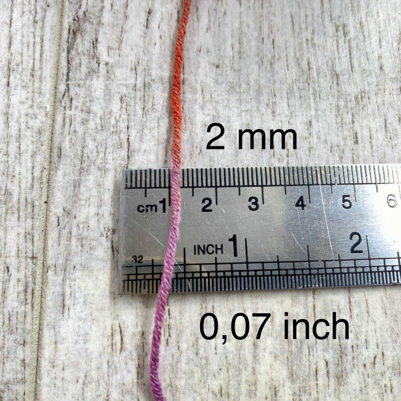 May include: A single strand of yarn with a color gradient transitioning from orange to pink. A metal ruler is positioned horizontally across the yarn, showing a thickness of 2 mm and 0.07 inches. The yarn is placed vertically against the ruler.