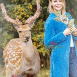 May include: A bright blue long-sleeved cardigan with a textured surface, worn over a white top and dark pinstriped trousers. The garment features a straight hemline and a simple open front design, providing a solid color contrast to the natural surroundings.