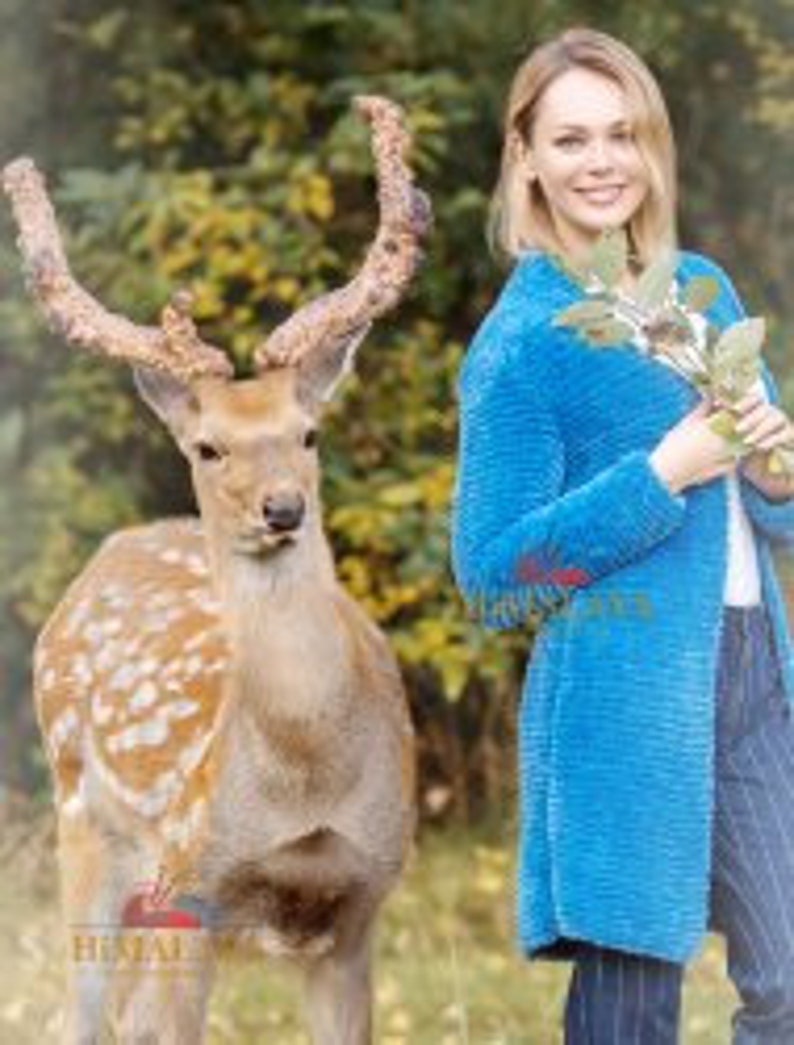 May include: A bright blue long-sleeved cardigan with a textured surface, worn over a white top and dark pinstriped trousers. The garment features a straight hemline and a simple open front design, providing a solid color contrast to the natural surroundings.