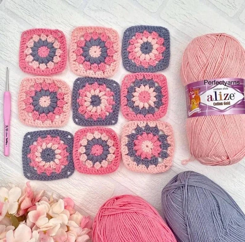 May include: Nine square crochet motifs featuring a central flower pattern in shades of pink, light pink, and grey. A pink crochet hook, a ball of pink Alize Cotton Gold yarn with a label, and two additional skeins of pink and grey yarn.