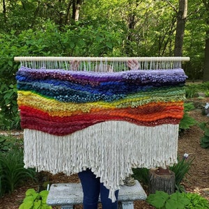 Pu&ograve; includere: Un grande arazzo da parete con un design paesaggistico orizzontale caratterizzato da strati di filato spesso e testurizzato nelle tonalit&agrave; viola, blu, verde, giallo, arancione e rosso, appeso a un bastone di legno con lunghe frange color crema.