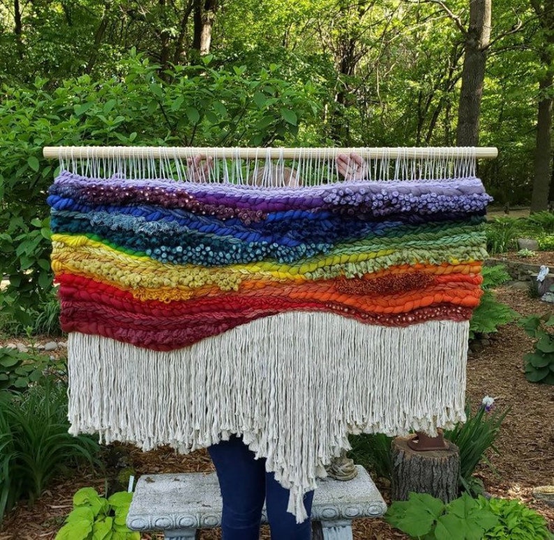 Pu&ograve; includere: Un grande arazzo da parete con un design paesaggistico orizzontale caratterizzato da strati di filato spesso e testurizzato nelle tonalit&agrave; viola, blu, verde, giallo, arancione e rosso, appeso a un bastone di legno con lunghe frange color crema.