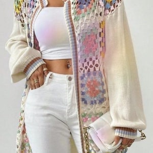 May include: A long-sleeved open-front cardigan featuring a cream-colored knit base with multicolored crochet granny square panels, paired with a white tube top, white jeans, and a white quilted handbag with a gold chain strap.