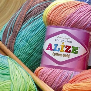 May include: A skein of Alize Cotton Gold yarn featuring a multi-colored gradient of yellow, pink, and orange. The label displays the text Batch Design cotton club ALIZE Cotton Gold. Additional skeins in blue, brown, and green are visible nearby.