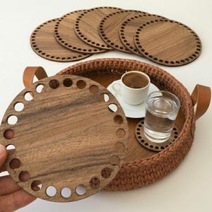 May include: A round wooden base with a series of holes along the perimeter, held in front of a larger round tray with a brown crocheted rim and leather handles. The tray contains a white coffee cup on a saucer and a clear glass of water.
