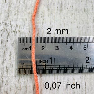 May include: A single strand of peach-colored yarn positioned vertically against a metal ruler. The ruler shows the yarn has a diameter of 2 mm or 0.07 inches. The yarn is a solid, matte, light orange color with a visible twisted fiber texture.