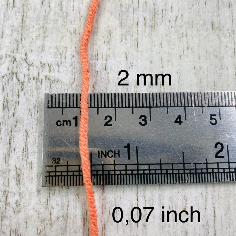 May include: A single strand of peach-colored yarn positioned vertically against a metal ruler. The ruler shows the yarn has a diameter of 2 mm or 0.07 inches. The yarn is a solid, matte, light orange color with a visible twisted fiber texture.