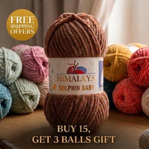 May include: A brown Himalaya Dolphin Baby yarn ball is the focal point, with a label featuring a logo. Other yarn balls in various colors, including pink, green, and red, are visible. The text "BUY 15, GET 3 BALLS GIFT" is also present.