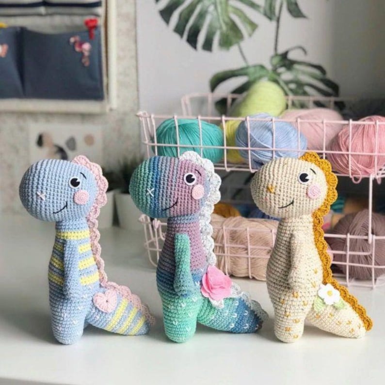May include: Three crocheted dinosaur toys standing side by side. The left one is light blue with yellow stripes and a pink heart. The middle one features a gradient of green, purple, and blue with a pink flower. The right one is cream with yellow accents and a white flower.