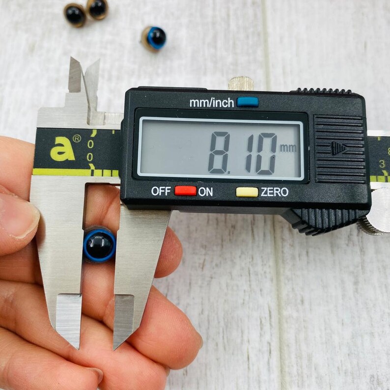 May include: A small round craft eye with a black pupil and a blue iris, held within the jaws of a digital caliper. The digital display on the black and silver measuring tool clearly shows a measurement of 8.10 mm for the diameter of the eye.