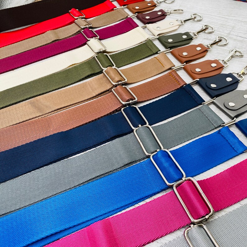 May include: A collection of adjustable bag straps in various solid colors including red, burgundy, cream, olive green, tan, brown, navy blue, grey, royal blue, and bright pink. Each strap features silver-toned metal hardware and leather-like tabs.