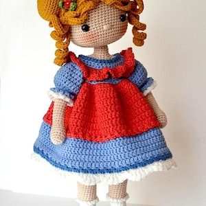 May include: A handmade crochet doll with golden yarn hair styled in two buns, featuring small green and red floral accents. The doll wears a blue dress with a red bodice overlay, white ruffled trim at the hem, white socks, and blue shoes.