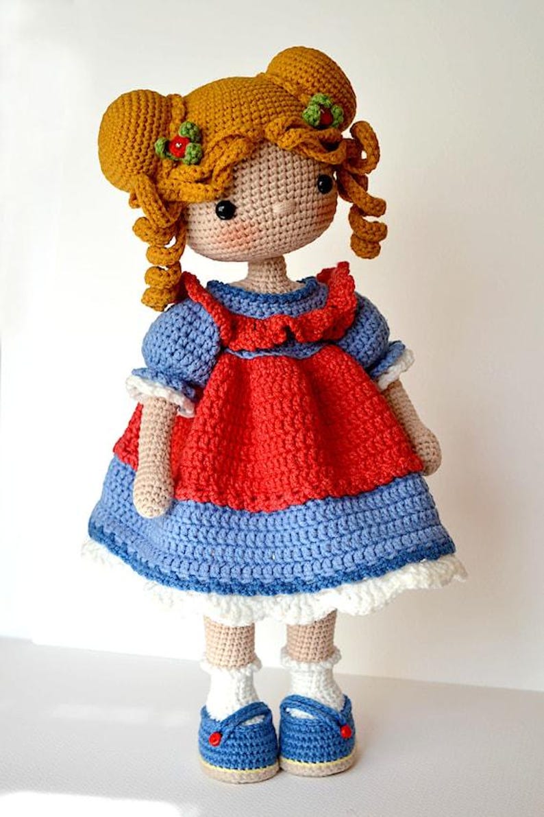 May include: A handmade crochet doll with golden yarn hair styled in two buns, featuring small green and red floral accents. The doll wears a blue dress with a red bodice overlay, white ruffled trim at the hem, white socks, and blue shoes.
