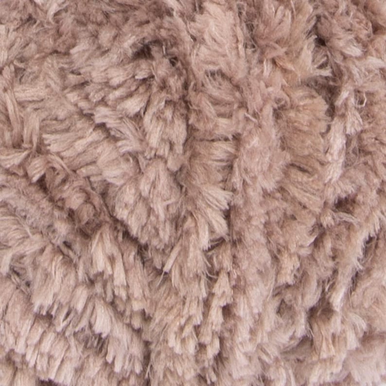 May include: A close-up view of a ball of yarn featuring a soft, shaggy, and textured appearance. The yarn is a solid, muted taupe or light brownish-gray color, characterized by numerous fine, fuzzy fibers that create a dense and plush surface.
