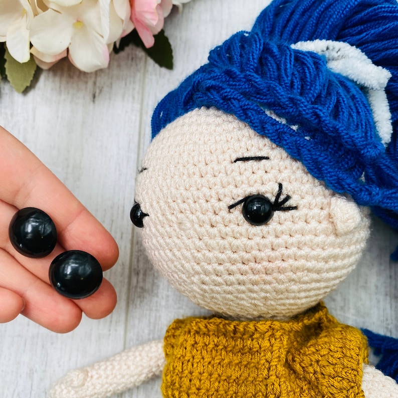 May include: A crocheted doll with beige skin, a mustard yellow top, and vibrant blue yarn hair featuring a white accent. The doll has black plastic safety eyes and embroidered eyelashes. Two additional loose black plastic safety eyes are shown.