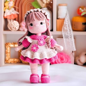 May include: A handmade crochet doll with brown yarn hair, wearing a white and pink layered dress adorned with two pink floral accents. The doll features a ruffled headband, rosy cheeks, and matching pink shoes, standing upright on a flat surface.