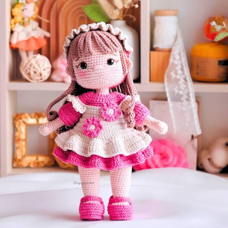 May include: A handmade crochet doll with brown yarn hair, wearing a white and pink layered dress adorned with two pink floral accents. The doll features a ruffled headband, rosy cheeks, and matching pink shoes, standing upright on a flat surface.