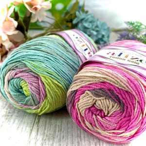 May include: Two skeins of Alize Cotton Gold yarn. The left skein features a variegated blend of teal, lime green, and soft lavender. The right skein displays a gradient of vibrant magenta, soft pink, and beige tones, each with a paper label.