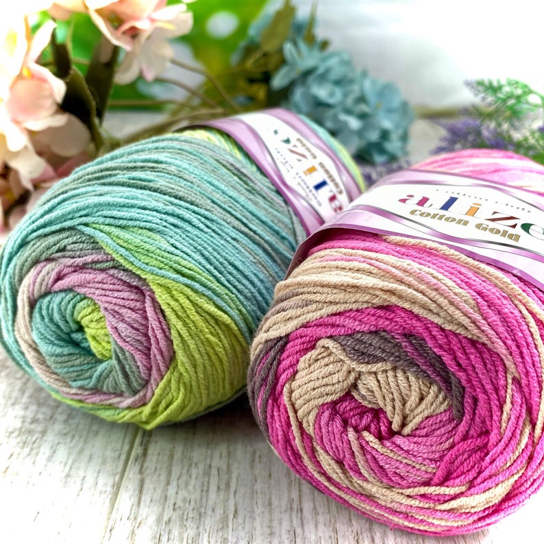 May include: Two skeins of Alize Cotton Gold yarn. The left skein features a variegated blend of teal, lime green, and soft lavender. The right skein displays a gradient of vibrant magenta, soft pink, and beige tones, each with a paper label.