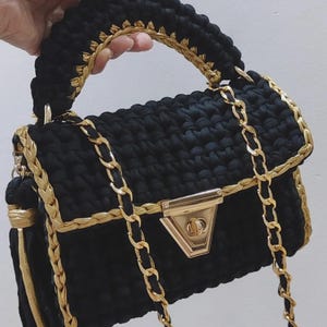 May include: A black crochet handbag with gold chain straps and trim. The bag has a gold clasp and a braided handle. A gold tassel hangs from the side, adding a decorative element to this stylish accessory.