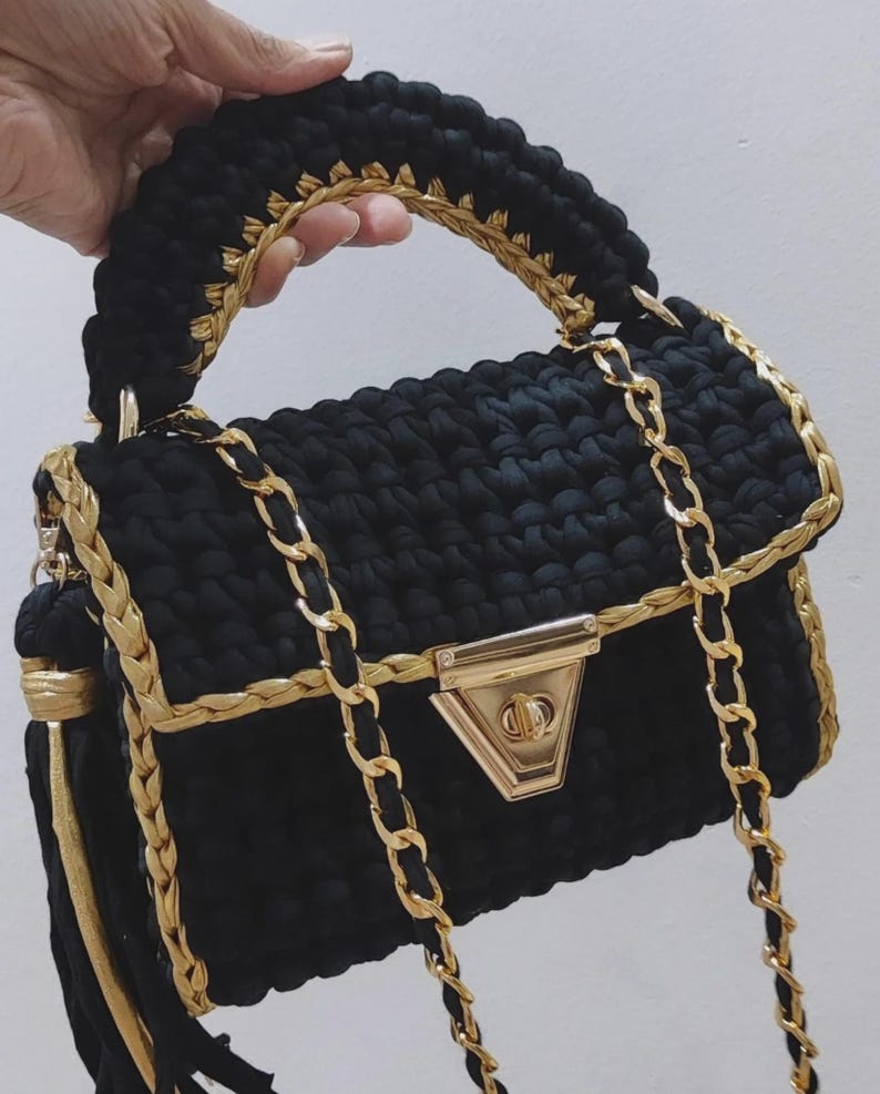 May include: A black crochet handbag with gold chain straps and trim. The bag has a gold clasp and a braided handle. A gold tassel hangs from the side, adding a decorative element to this stylish accessory.