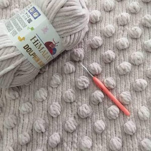 May include: A close-up of a light gray yarn ball with a label that reads "Himalaya Dolphin Baby". A pink crochet hook rests on a textured, light gray fabric with raised, round, pom-pom-like details. The yarn is soft and fluffy.