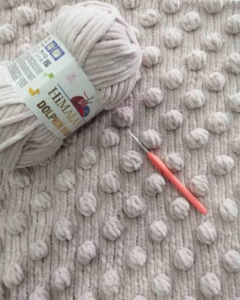 May include: A close-up of a light gray yarn ball with a label that reads "Himalaya Dolphin Baby". A pink crochet hook rests on a textured, light gray fabric with raised, round, pom-pom-like details. The yarn is soft and fluffy.