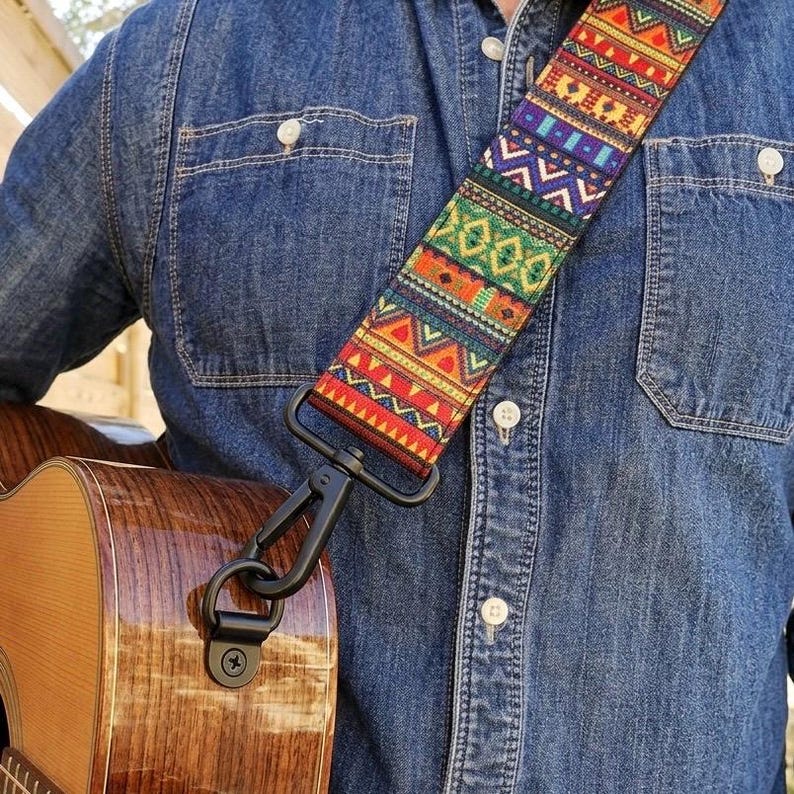 May include: A guitar strap featuring a vibrant, multi-colored woven pattern with geometric shapes in red, orange, yellow, green, blue, and purple. The strap is attached to a wooden acoustic guitar with a black metal clip and hardware.