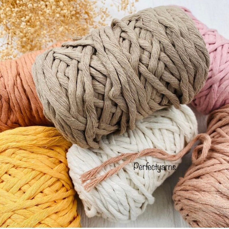 Pu&ograve; includere: Una collezione di matasse di filato di cotone intrecciato spesso in vari colori, tra cui beige, giallo senape, crema e rosa polvere. La matassa color crema presenta il testo Perfectyarns stampato in nero sulla sua superficie.