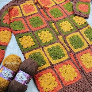 May include: A handmade crochet cardigan constructed from square motifs in shades of orange, yellow, olive green, and brown. Two skeins of yarn, one mustard yellow and one dark brown, are positioned at the bottom left of the garment.