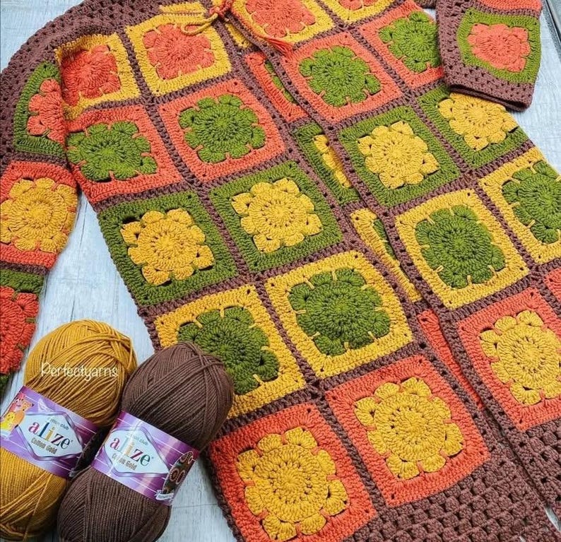 May include: A handmade crochet cardigan constructed from square motifs in shades of orange, yellow, olive green, and brown. Two skeins of yarn, one mustard yellow and one dark brown, are positioned at the bottom left of the garment.
