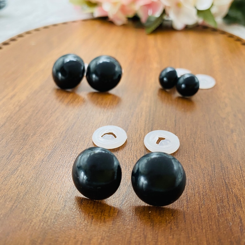May include: A collection of six smooth, spherical black plastic safety eyes in varying sizes, each paired with a translucent white plastic washer backing, arranged in three distinct pairs on a smooth, light brown wooden surface.