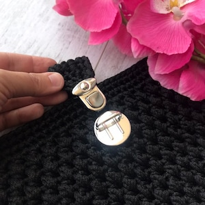 May include: A close-up shot of black crocheted fabric with a silver metal clasp. The clasp is open, showing the mechanism. A hand holds the fabric. Pink flowers are in the background.