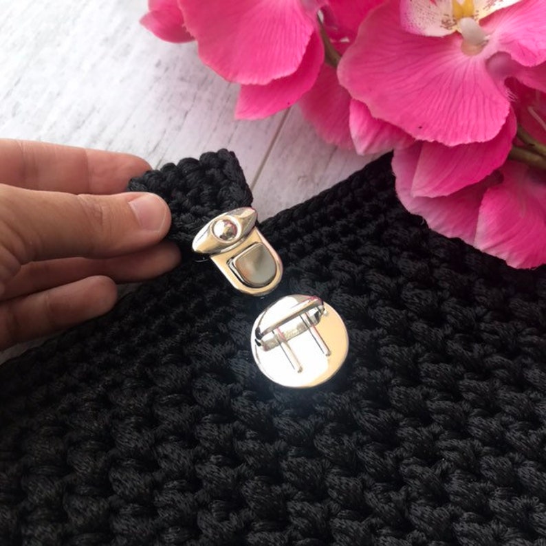 May include: A close-up shot of black crocheted fabric with a silver metal clasp. The clasp is open, showing the mechanism. A hand holds the fabric. Pink flowers are in the background.