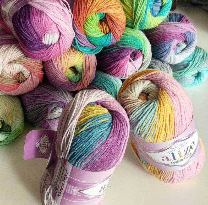 May include: A collection of multi-colored yarn skeins featuring variegated gradients of pink, purple, yellow, green, and blue. Each skein is wrapped in a paper label with the text Alize Cotton Gold and &Ouml;rmeye Değer printed on the front.