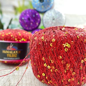 May include: A ball of bright red yarn interwoven with small, reflective gold sequins. A black paper label is wrapped around the yarn, featuring the text Himalaya Yildiz in gold lettering and a small graphic of knitting needles and yarn.