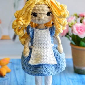 May include: A handcrafted crochet doll featuring long, curly yellow yarn hair, a black headband with a bow, and a light blue dress with a white apron. The doll has dark eyes, white tights, and black shoes, all constructed from textured yarn.