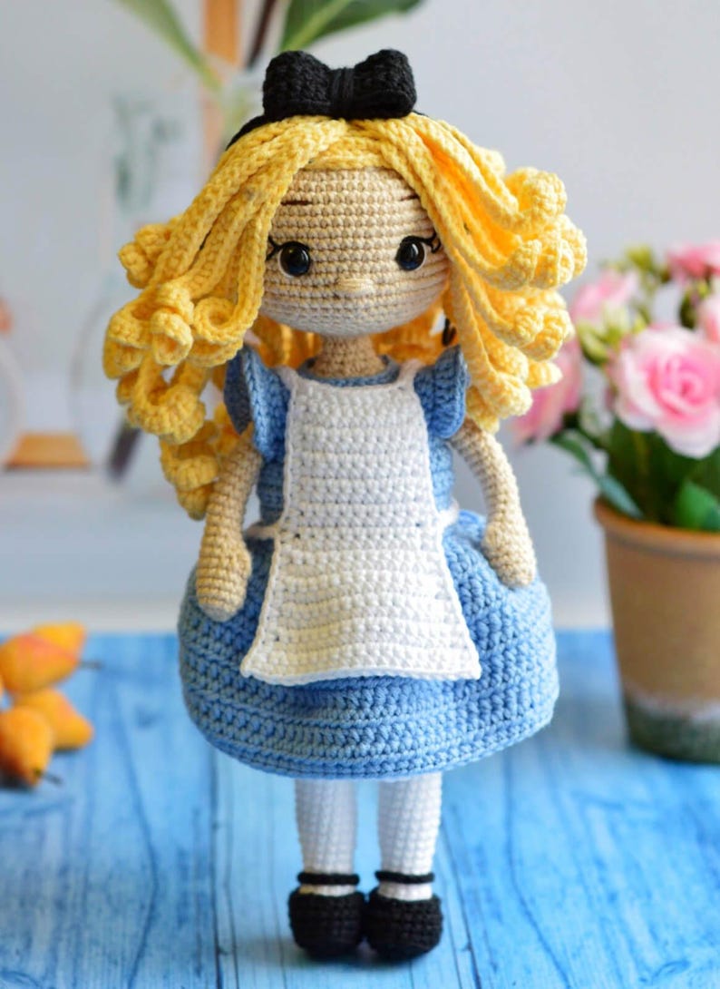 May include: A handcrafted crochet doll featuring long, curly yellow yarn hair, a black headband with a bow, and a light blue dress with a white apron. The doll has dark eyes, white tights, and black shoes, all constructed from textured yarn.