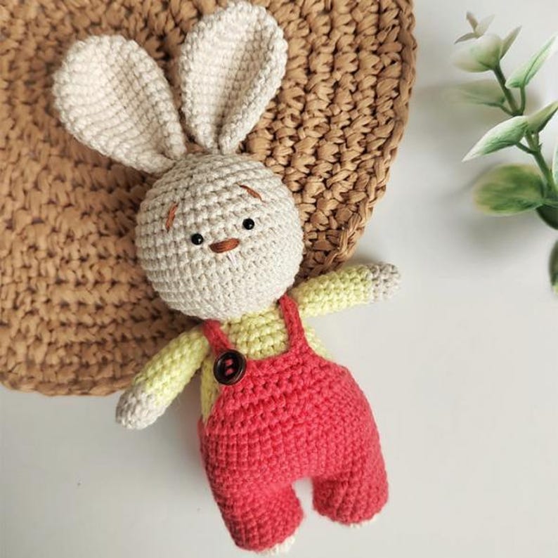 May include: A crochet rabbit doll with long ears, a cream-colored head, and embroidered facial features. The doll wears a pale yellow long-sleeved shirt under red overalls with a single dark brown button on the front chest strap area.