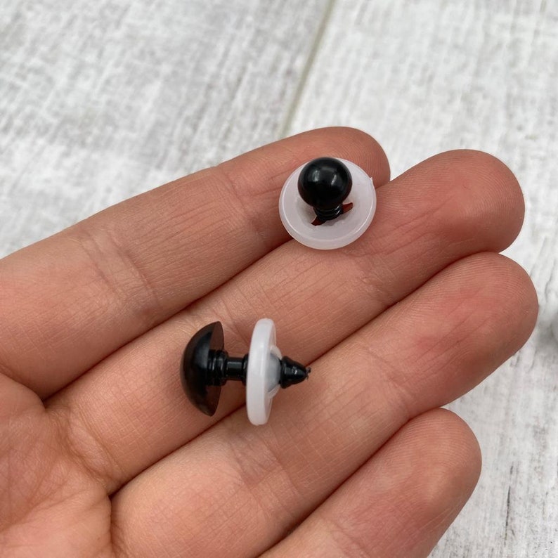 May include: Two black plastic safety eyes for plush toys, each featuring a smooth, rounded dome front and a threaded post with a translucent white plastic washer attached to secure the eye in place on a fabric surface.