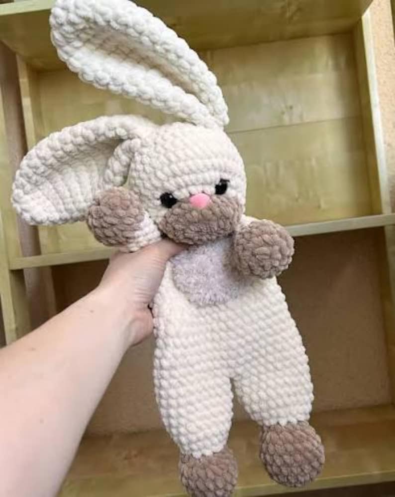 May include: A handmade, plush bunny toy in cream, brown, and pink. The bunny has long, floppy ears, a pink nose, and black eyes. The toy is made of a soft, textured material, and the arms and feet are a darker brown color.