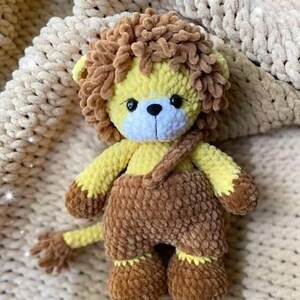 May include: A handmade crochet lion stuffed animal. The lion has a yellow body, brown mane, and brown overalls. The face is yellow and blue with black eyes and a black nose. The lion is sitting on a cream-colored blanket.